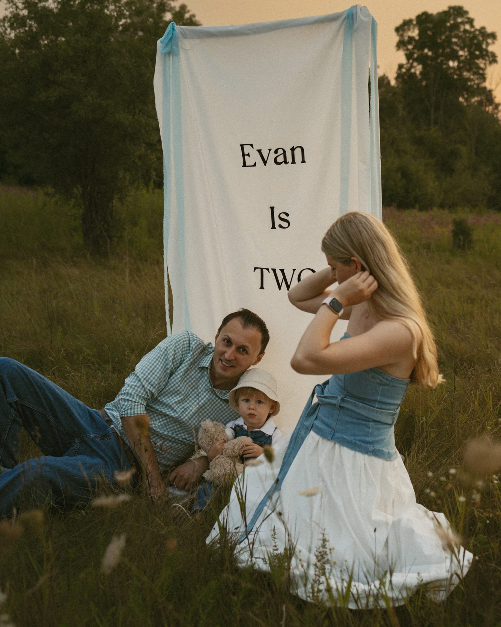 A mother, father and toddler sitting in a wildflower meadow beside a hanging fabric banner at sunset