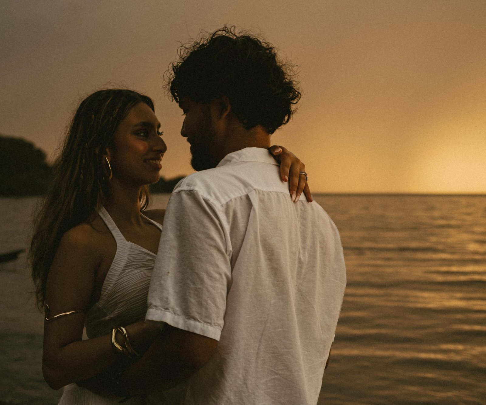 A couple embracing on a lakeshore in warm sunset light, the sky glowing orange behind them