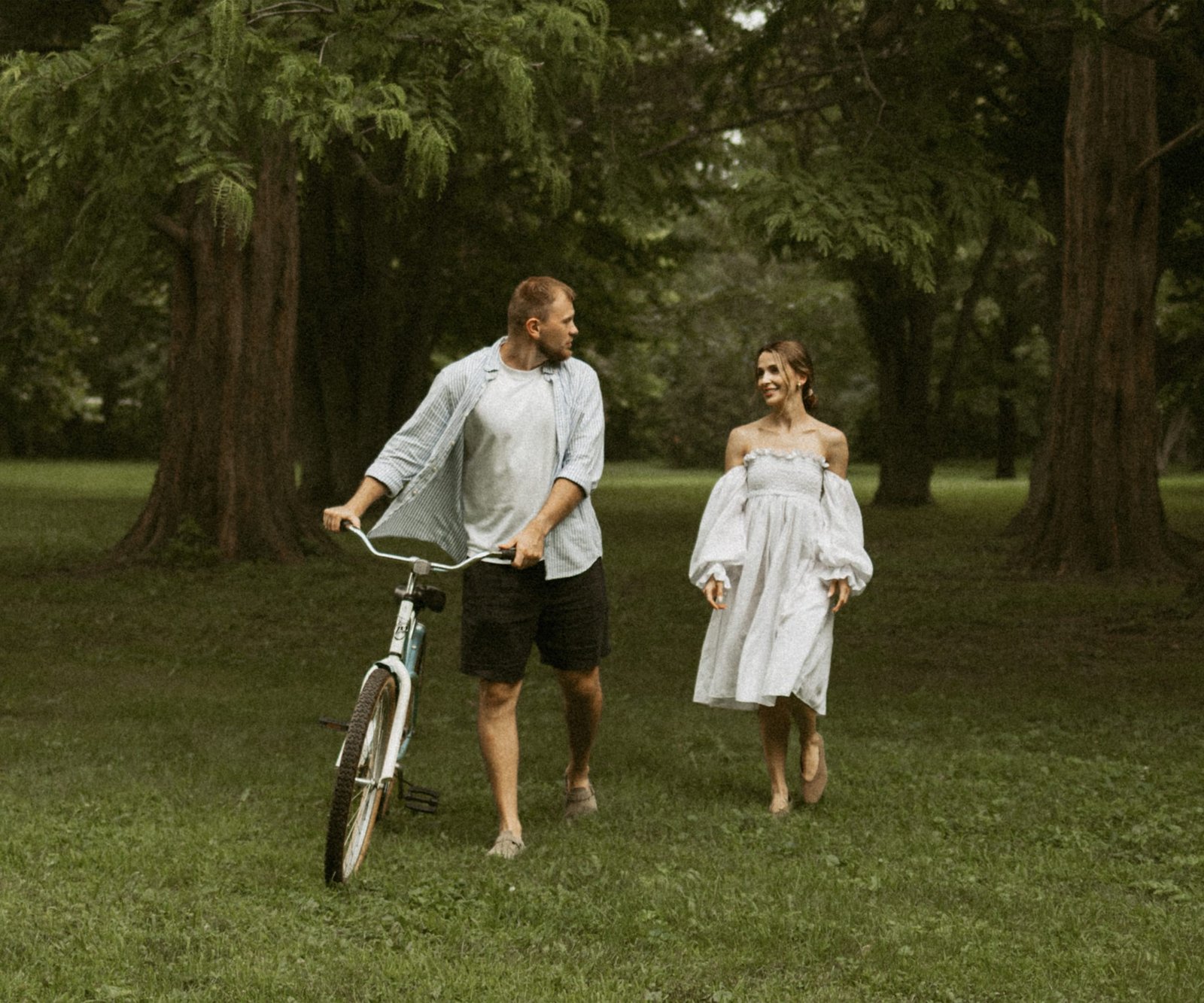 A couple walking together across a park lawn, the man wheeling a vintage bicycle beside her