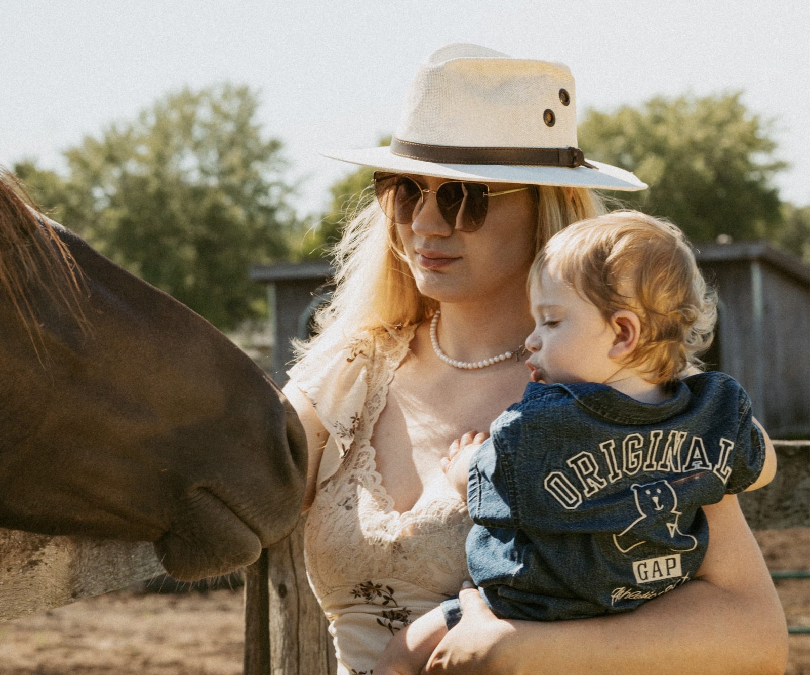 A mother in a wide-brim hat holding her toddler as a horse nuzzles close at a paddock fence